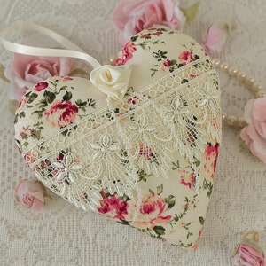 May include: A heart-shaped pillow with a floral print and lace trim. The pillow is cream-colored with pink and red roses. It is surrounded by pink roses and a pearl necklace.