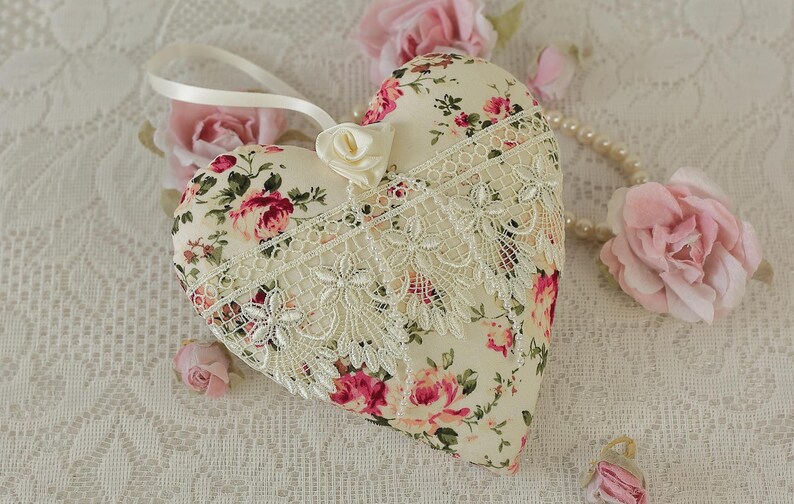 May include: A heart-shaped pillow with a floral print and lace trim. The pillow is cream-colored with pink and red roses. It is surrounded by pink roses and a pearl necklace.
