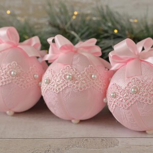 May include: Three pink Christmas baubles decorated with white lace and pearl accents. Each bauble has a pink satin ribbon bow.