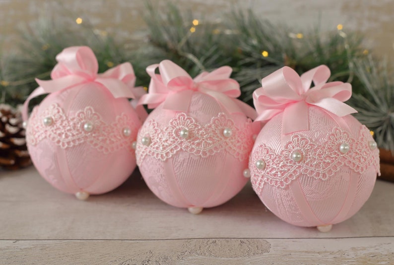 May include: Three pink Christmas baubles decorated with white lace and pearl accents. Each bauble has a pink satin ribbon bow.