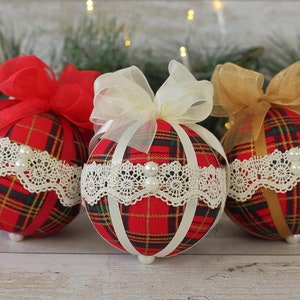 May include: Three red, white, and gold plaid Christmas ornaments with white lace trim and pearl accents. Each ornament has a different colored ribbon bow.