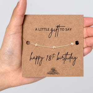 May include: A gold-toned chain bracelet featuring three small star charms, displayed on a rectangular brown card. The card text reads: A LITTLE gift TO SAY happy 18th birthday, with the brand name Swanky Crafts printed at the bottom.