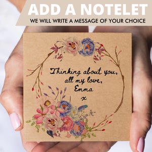 May include: A square kraft paper notelet featuring a watercolor floral wreath in shades of pink, blue, and green. The center contains the handwritten black text: Thinking about you, all my love, Emma x. The top banner reads ADD A NOTELET.
