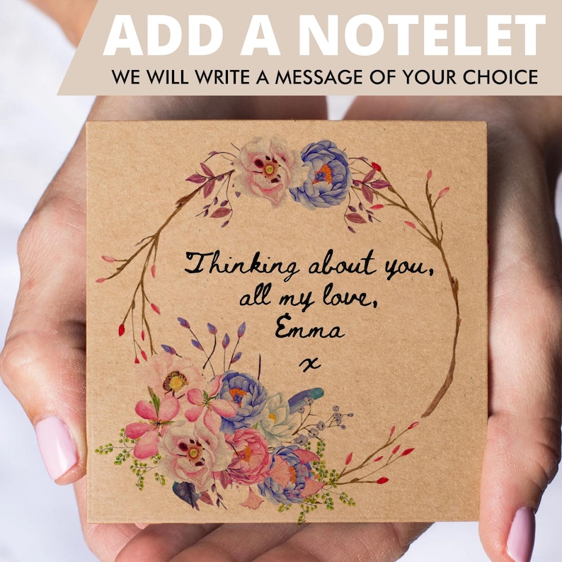 May include: A square kraft paper notelet featuring a watercolor floral wreath in shades of pink, blue, and green. The center contains the handwritten black text: Thinking about you, all my love, Emma x. The top banner reads ADD A NOTELET.