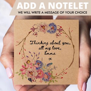 K&ouml;nnte beinhalten: Eine quadratische Notizkarte aus Kraftpapier mit einem Aquarell-Blumenkranz in Rosa-, Blau- und Gr&uuml;nt&ouml;nen. In der Mitte steht der handgeschriebene schwarze Text: Thinking about you, all my love, Emma x. Das obere Banner zeigt ADD A NOTELET.