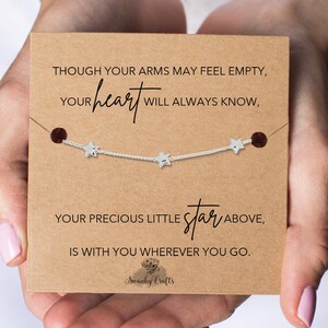 May include: A silver necklace with three star charms is presented on a brown card. The card features the text: "Though your arms may feel empty, your heart will always know, your precious little star above, is with you wherever you go."