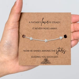 May include: A silver-toned chain bracelet featuring three small star charms, displayed on a rectangular brown card. The card includes the printed text: A FATHER'S love IS STEADY, IT NEVER FADES AWAY. NOW HE SHINES AMONG THE stars. GUIDING YOU EACH DAY.
