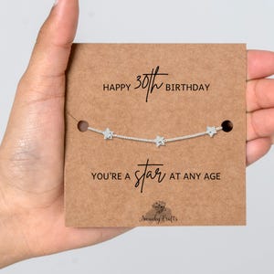 May include: A silver-toned chain bracelet featuring three small star charms, displayed on a rectangular brown card. The card text reads: HAPPY 30TH BIRTHDAY and YOU'RE A STAR AT ANY AGE, with the brand name Swanky Crafts printed at the bottom.