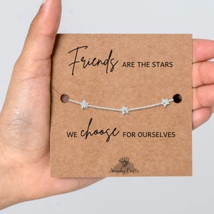 May include: A silver-toned chain bracelet featuring three small star charms, displayed on a rectangular brown card. The card text reads: Friends ARE THE STARS WE choose FOR OURSELVES, with the Swanky Crafts logo printed at the bottom.