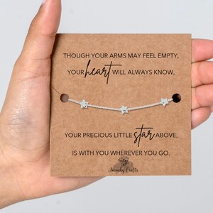 May include: A silver necklace with three star charms is shown on a brown card. The card has text that reads "Though your arms may feel empty, your heart will always know, your precious little star above, is with you wherever you go."