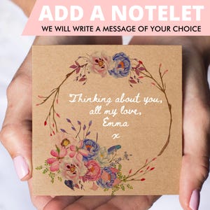 May include: A square kraft paper notelet with a floral wreath design. The wreath features pink, blue, and white flowers. The text "Thinking about you, all my love, Emma x" is written in white script. The top banner reads "ADD A NOTELET".