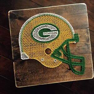 May include: Green Bay Packers football helmet made with string art on a wooden background. The helmet is green, yellow, and white.