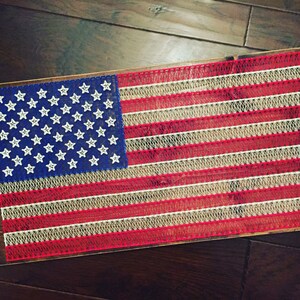 May include: A rectangular wooden board featuring a string art design of the American flag. The design uses blue string for the canton with white star-shaped nails, and alternating red and white string lines to represent the stripes of the flag.