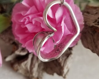 May include: A silver heart-shaped pendant necklace made from a repurposed spoon. The pendant is hanging from a silver chain and is resting on a pink flower.