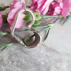 May include: A silver heart-shaped pendant necklace made from a repurposed spoon, hanging against a background of pink carnations.