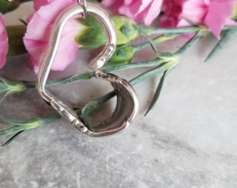 May include: A silver heart-shaped pendant necklace made from a repurposed spoon, hanging against a background of pink carnations.