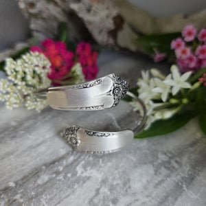 May include: A silver-toned bypass bracelet crafted from a repurposed spoon, featuring an open-ended design. The two ends of the bracelet display intricate floral embossing and decorative scrollwork patterns along the handle sections.