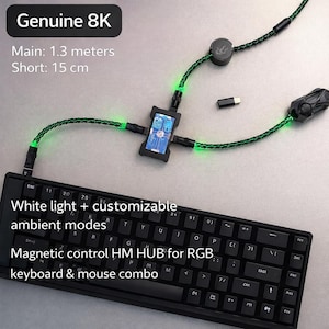 Pode incluir: Teclado preto com letras brancas, conectado a um hub preto com uma pequena tela e cabos verdes brilhantes. O texto "Genuine 8K" e "Magnetic control HM HUB for RGB keyboard & mouse combo" são visíveis.