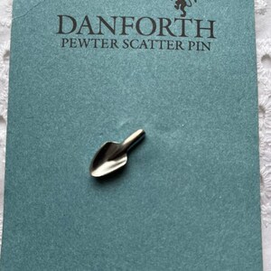 May include: A silver pewter scatter pin shaped like a small shovel. The pin is on a blue card with the text "DANFORTH PEWTER SCATTER PIN" and "DANFORTH PEWTERERS LTD. MADE IN MIDDLEBURY, VERMONT USA".