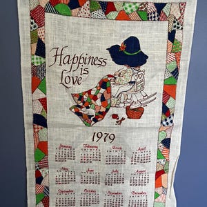 May include: A 1979 fabric calendar wall hanging featuring a printed illustration of a person in a blue hat sitting in a rocking chair with a patchwork quilt. The border consists of colourful patchwork triangles. The text reads Happiness is Love.
