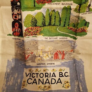 May include: A vintage linen tea towel with a colorful illustration of Victoria, British Columbia, Canada. The design features a totem pole, Parliament Buildings, the Butchart Gardens, and Undersea Gardens. The text "Victoria B.C. Canada" is printed in black on a blue background.