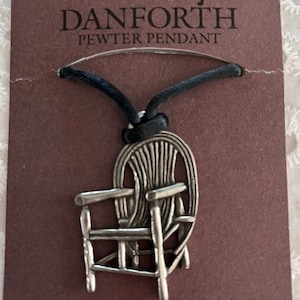 May include: A silver pewter pendant of a rocking chair on a black cord. The pendant is on a brown card with the text "DANFORTH PEWTER PENDANT" and "DANFORTH PEWTERERS LTD. MADE IN MIDDLEBURY, VERMONT USA".