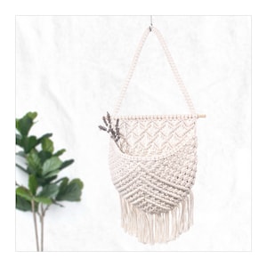 May include: A white macrame wall hanging with a wooden dowel and a pocket. The hanging has a fringe at the bottom and is decorated with dried lavender.