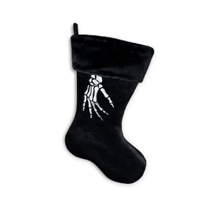 May include: Black Christmas stocking with a white skeleton hand design. The stocking has a plush, velvety texture and a folded-over cuff. A loop is attached for hanging. Perfect for Halloween or gothic decor.