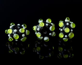 May include: Three black glass beads with white and lime green accents. The beads are round and have a textured surface. They are arranged in a row on a black background.