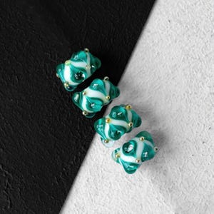 Puede incluir: Cuatro cuentas de vidrio de color verde azulado y blanco con detalles dorados. Las cuentas tienen un patrón de espiral y están dispuestas en una fila sobre una superficie blanca y negra.