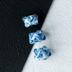 May include: Three handmade glass beads with a marbled blue and white design. Each bead has a unique shape and is accented with small silver-coloured metal embellishments. The beads are displayed against a black and white background.