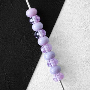 Puede incluir: Una hebra de cuentas de vidrio lavanda con lunares blancos. Las cuentas están enhebradas en un alambre plateado.