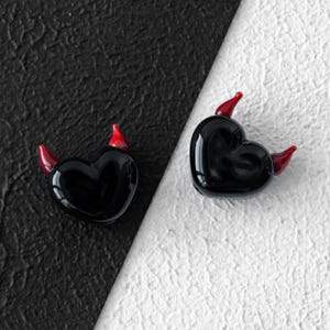 May include: Two black heart-shaped glass ornaments with red devil horns. The ornaments are displayed on a black and white textured background. The hearts are glossy and reflect light.