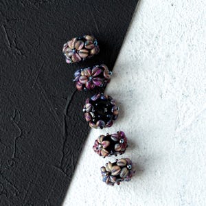 Puede incluir: Cinco cuentas florales hechas a mano en tonos morados, marrones y azules. Cada cuenta presenta un diseño floral con múltiples pétalos y pequeños acentos azules. Las cuentas están dispuestas verticalmente sobre un fondo blanco y negro.