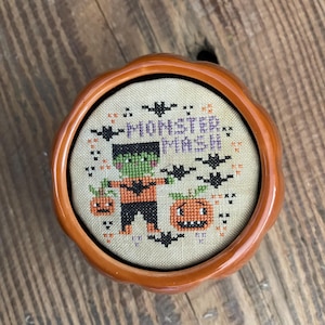 May include: A round orange ceramic pumpkin-shaped container holding a cross-stitch embroidery piece. The fabric features a green-skinned monster, two jack-o'-lanterns, and black bats with the text "MONSTER MASH" stitched in purple thread.