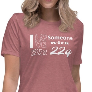 May include: A dusty rose t-shirt with the white text "I Love Someone with 22q". The text is arranged vertically with a heart symbol above the word "Love" and three heart symbols below it. The shirt is a relaxed fit.