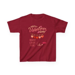 Könnte beinhalten: Burgunderfarbenes T-Shirt mit Valentinstags-Design. Das Design enthält den Text "Happy Valentines Day" und "Insert Candy Here". Darunter hängen Herzen und ein herzförmiger Pfeil.