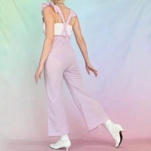 May include: A pink and white gingham jumpsuit with ruffled straps. The jumpsuit has a wide leg and is worn with white shoes.