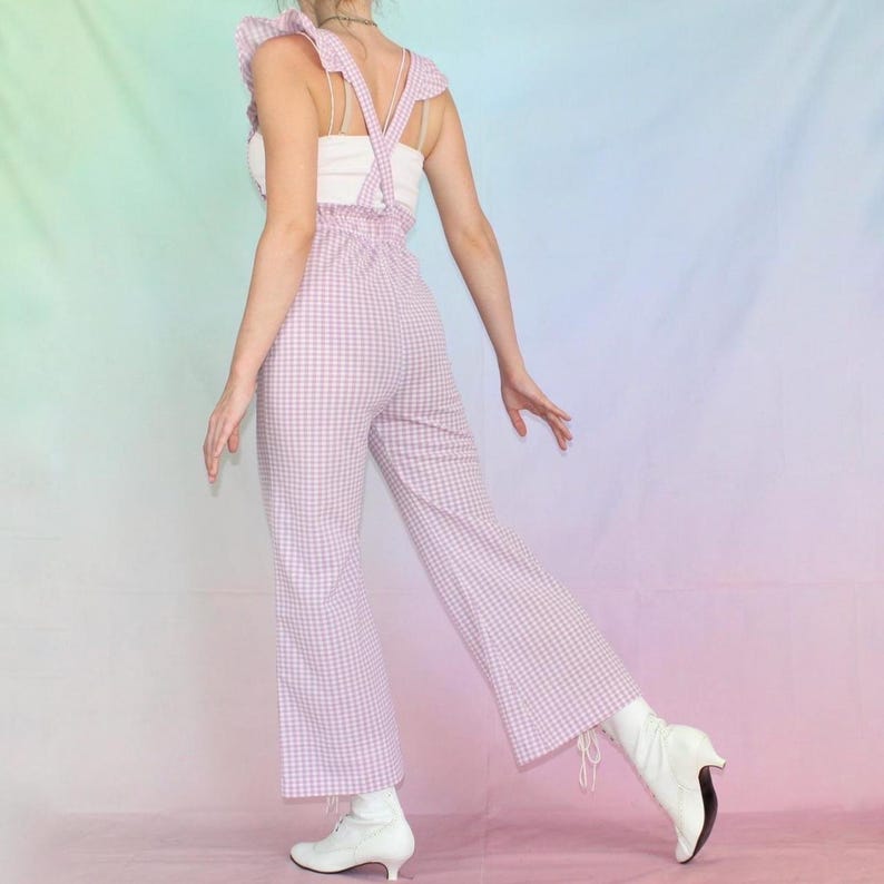 May include: A pink and white gingham jumpsuit with ruffled straps. The jumpsuit has a wide leg and is worn with white shoes.