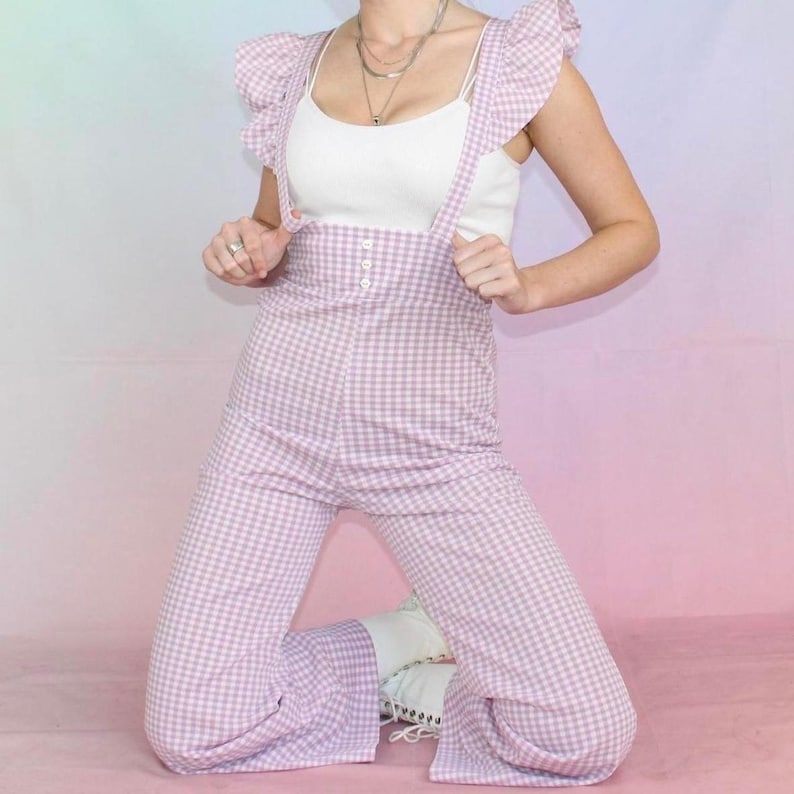 May include: A purple and white gingham jumpsuit with ruffled straps and a white button closure. The jumpsuit has a wide leg style.