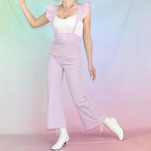 May include: A pink and white gingham jumpsuit with a white tank top underneath and white ruffle sleeves. The jumpsuit has suspenders and a button closure. The model is wearing white heeled boots.
