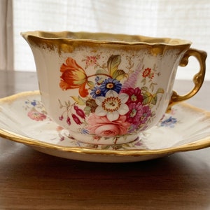 May include: A white teacup and saucer with a floral pattern in shades of pink, red, blue, and yellow. The cup and saucer have a gold rim.