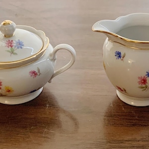 May include: A white porcelain sugar bowl with a lid and a creamer. Both pieces are decorated with small floral patterns in pink, blue, and yellow, and have gold trim. The sugar bowl has a small gold knob on the lid.