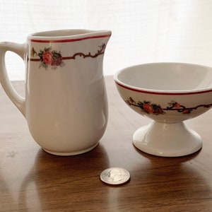 May include: A white ceramic creamer pitcher and a small pedestal bowl. Both pieces have a red trim and are decorated with a floral pattern of pink and purple roses. The creamer has a handle, and the bowl sits on a short, rounded base.