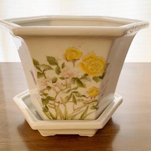 May include: A white ceramic planter with a matching saucer. The planter features a hand-painted floral design with yellow roses, pink flowers, and green leaves. The planter has a unique octagonal shape.