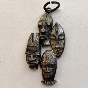 May include: A silver pendant featuring four stylized African masks. The masks are arranged in a cluster and have a textured, tribal design.