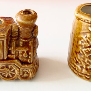May include: Two small ceramic figurines, one shaped like a train engine and the other like a mug with a windmill design. Both are brown and have a rustic, vintage look.