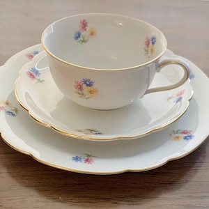 May include: A white porcelain teacup, saucer, and plate set. The set features a gold trim and is decorated with small floral patterns in pink, blue, and yellow. The edges of the plate and saucer have a scalloped design.