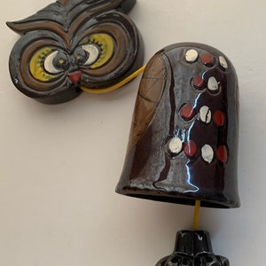 May include: A ceramic owl wind chime with a brown, black, and white painted design. The owl has large, yellow eyes and a small, red beak. The chime is attached to a yellow string.