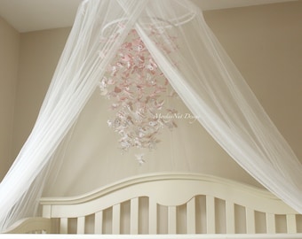 May include: A white canopy hangs over a white crib with a pink and white butterfly mobile hanging from the ceiling. The mobile has the text "Hoosier Nest Designs" on it.